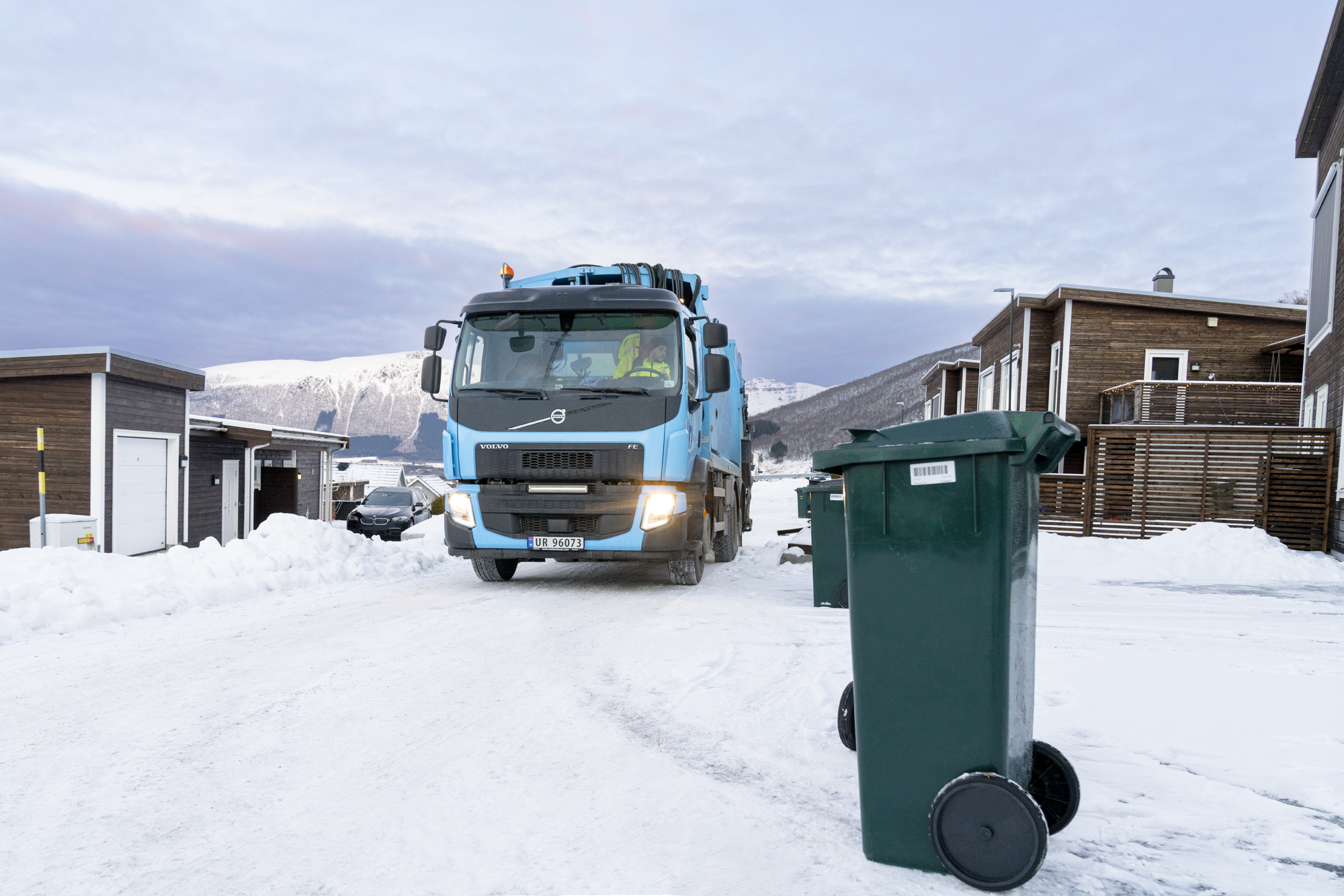 Renovasjonsbil på vei inn i boligområde, med en avfallsdunk i forgrunnen.