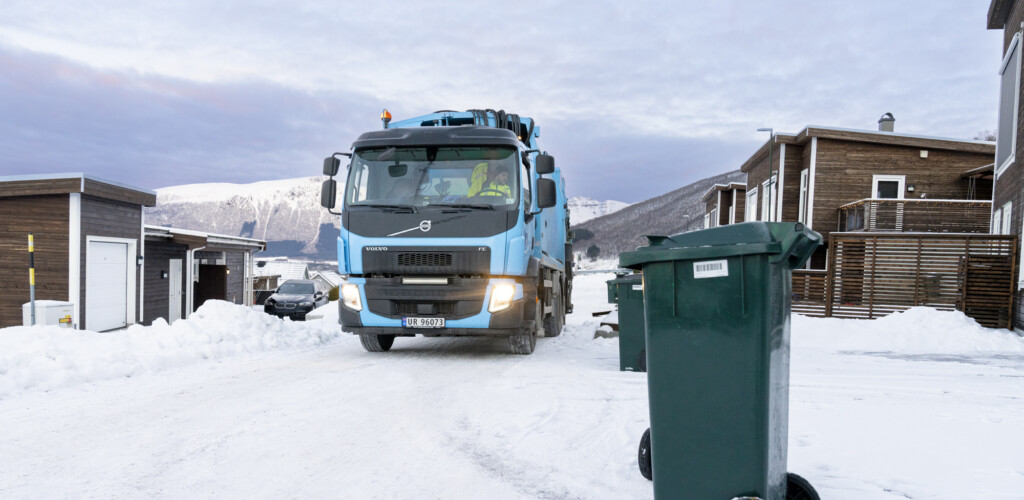 Renovasjonsbil på vei inn i boligområde, med en avfallsdunk i forgrunnen.